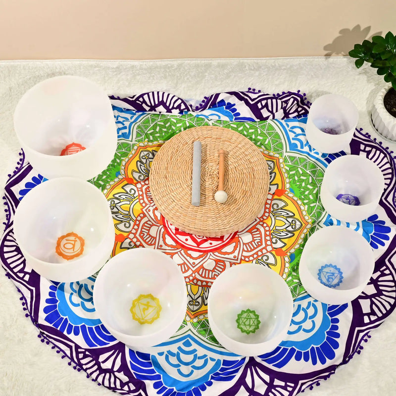 Set of seven white crystal chakra bowls with colored symbols, arranged around a woven mat.