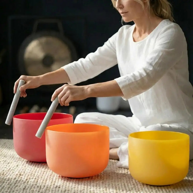 Three vibrant, translucent singing bowls in pink, orange, and yellow sit on a textured surface.