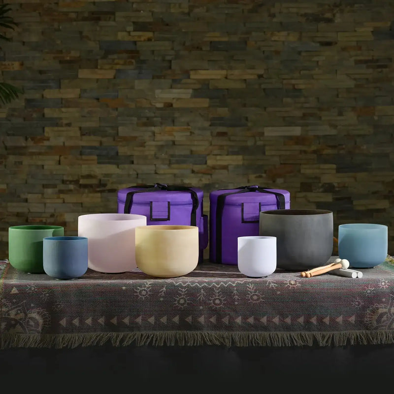 Set of seven colorful, handcrafted crystal singing bowls in various sizes and hues including green, blue, white, yellow, gray, and teal, displayed with their purple carrying cases.