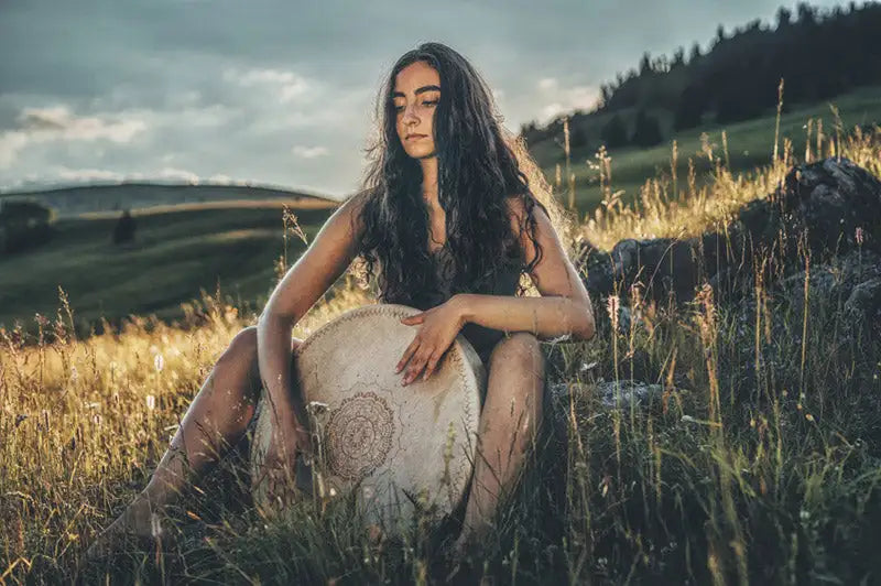 A cream-colored, handcrafted drum with intricate embroidered patterns rests in the grass beside a seated woman.