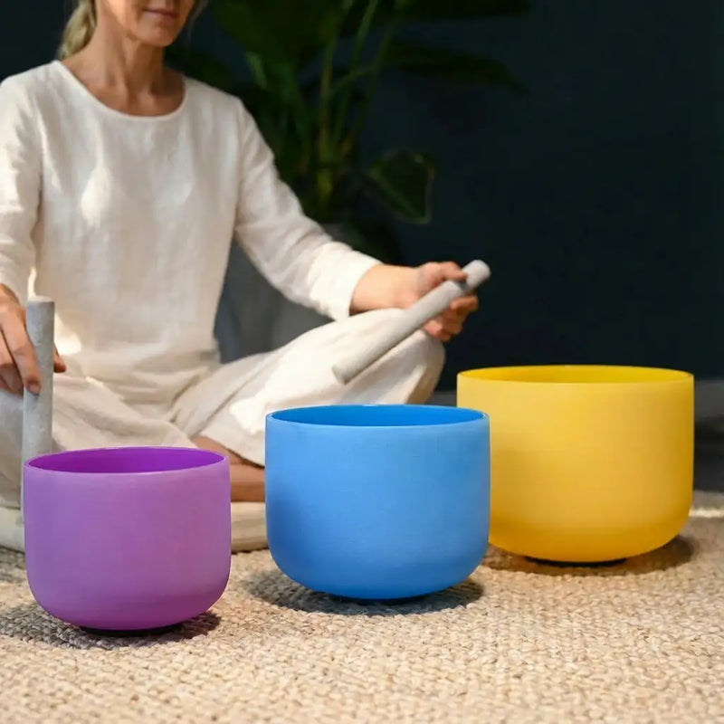 Three vibrant, matte-finished crystal singing bowls in purple, blue, and yellow are arranged on a textured surface.