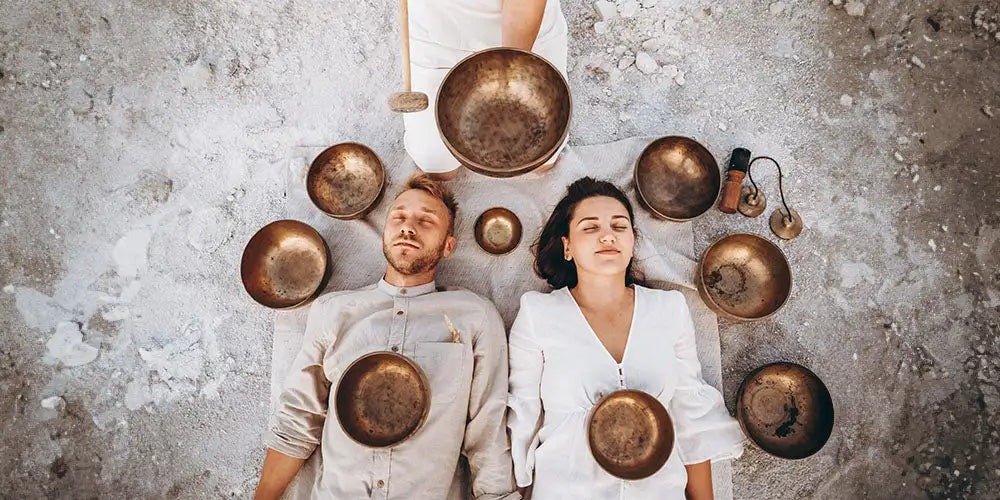 A set of bronze-toned singing bowls arranged around a man and woman lying on their backs.